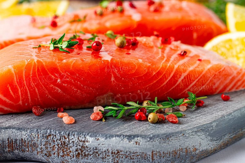 Two pieces of salmon fillet sit on a wooden cutting board. They are topped with herbs and pink peppercorns. Slices of lemon and lime surround the fish, creating a vibrant setting for cooking