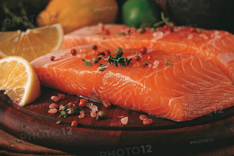Two salmon fillets rest on a wooden platter. The fillets are garnished with herbs and spices. Lemon and lime slices are nearby. This setup is ready for preparation in a kitchen