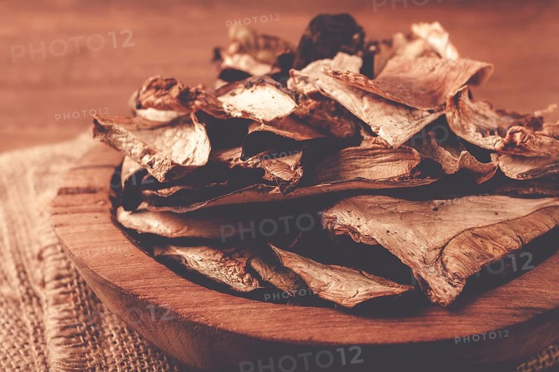 A collection of dried mushrooms fills a wooden bowl placed on a cloth surface. The mushrooms are cut into thin slices, ideal for adding to various dishes