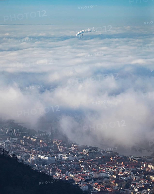 Aerial view of the city of Heidelberg on the Neckar, sea of fog from low-hanging clouds and fog fields, sunlit above the roofs of the city and the Neckar river, floating and waving with large and smoking chimney vents sticking out of the fog in the background, Heidelberg, Baden-Württemberg, Germany