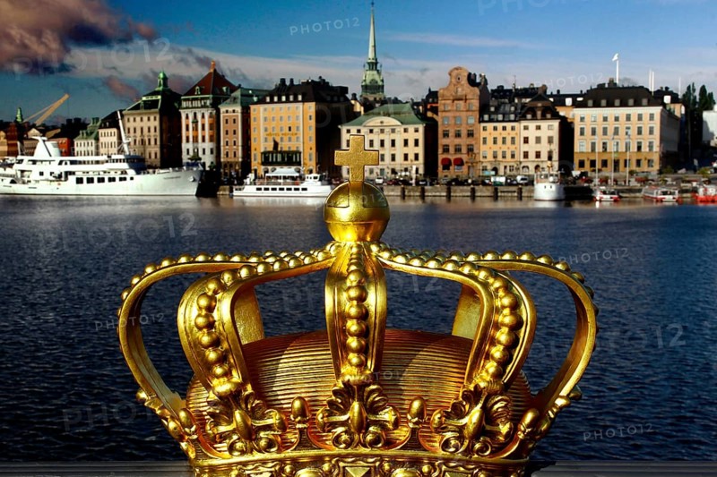 A golden crown in the foreground with a view of the skyline and boats of Stockholm, Stockholm, Gamla Stan, Sweden