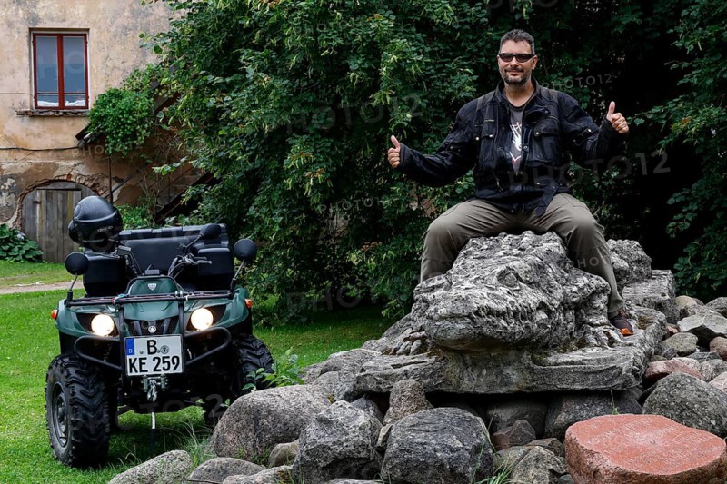 Person sitting on a crocodile sculpture next to an ATV in a green setting, Dundaga, Latvia