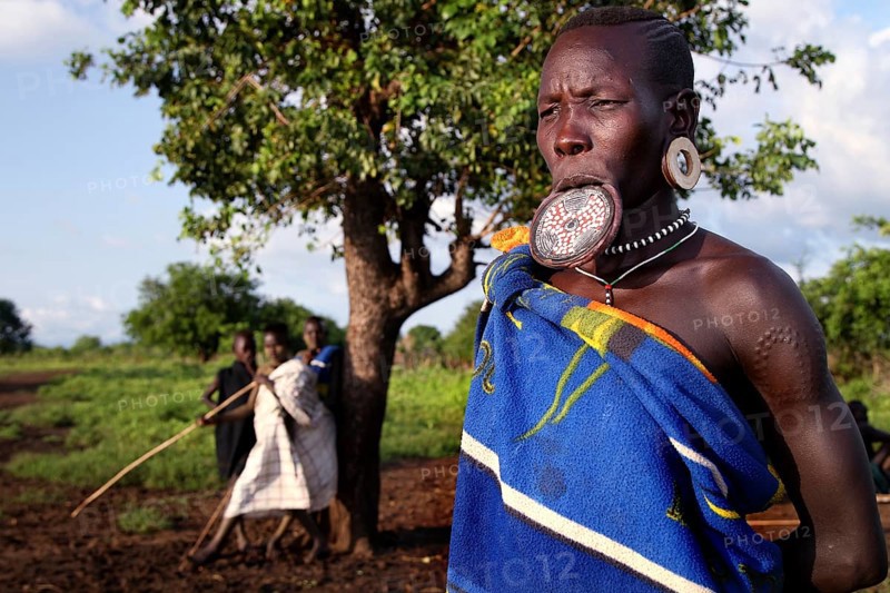 Mursi woman with lip plate in front of a tree in Mago National Park. A village can be seen in the background, Mago National Park, Ethiopia