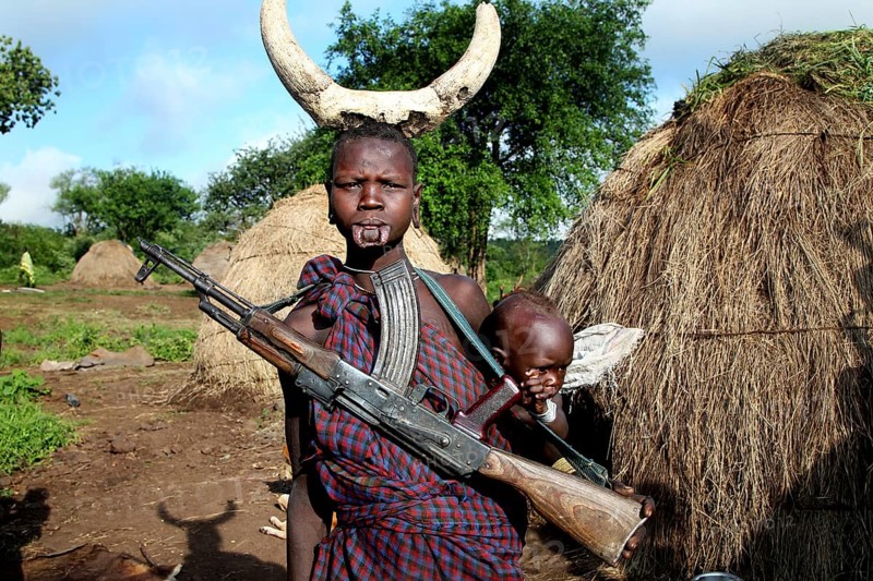 Man with gun and child on his back, straw huts in the background in Mago National Park, Mago National Park, Ethiopia