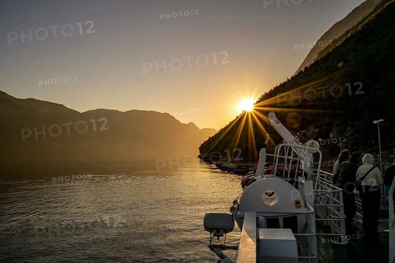 A boat sails through the Lysefjord at sunset with sun star with calm water and surrounded by mountains, view from the Columbus ferry, Lysefjord, Rogaland, Norway