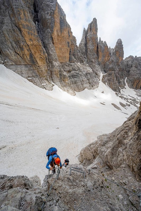 Mountaineer climbs a ladder on the Via Ferrata dell'Ideale via ferrata, spectacular mountain landscape with steep rock faces, remains of the Vedretta d'Ambiez glacier in the back, Brenta Mountains, Parco Naturale Brenta-Adamello, Trentino, Italy
