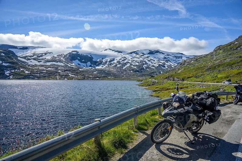 Geiranger, Møre og Romsdal Province, Norway, BMW G 650 GS Sertao Enduro motorcycle trip along a lake with snow-covered mountains under a blue sky