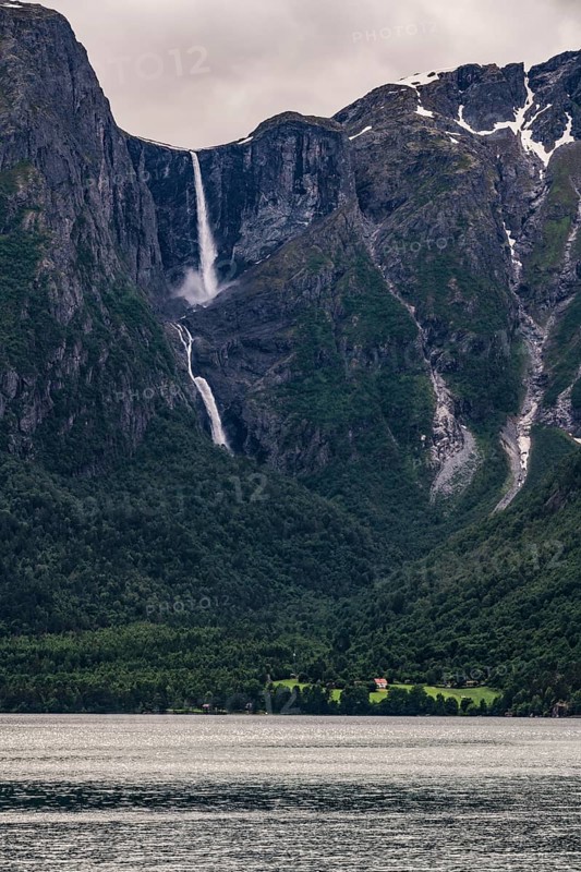 A narrow waterfall plunges from dramatic mountains into a dark fjord