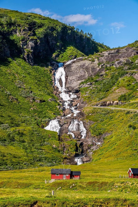 A cascading waterfall flows over green hills next to a red hut in the sunshine