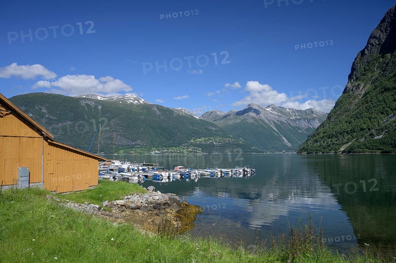 Norddal, Møre og Romsdal province, Norway, wooden cabin on the shore of a fjord with boats and mountains in the background under blue sky