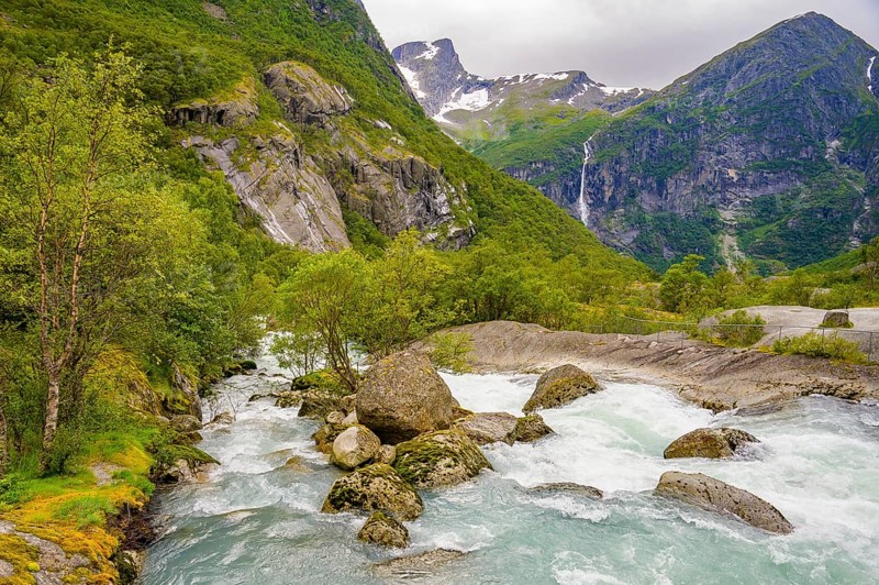 Briksdalsbre, Vestland, Norway, fast-flowing river through green valley with rocks and distant waterfalls on Brikdalsbreen glacier