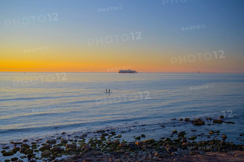 Hirtshals, Nordjylland region, Denmark, sunset over a calm sea with gentle waves and rocks on the shore in the background a ferry from Hirtshals to Kristiansand in the foreground two stand-up paddlers