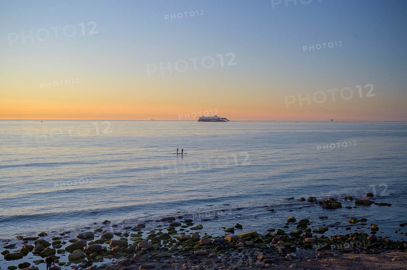 Hirtshals, Nordjylland region, Denmark, sunset over a calm sea with gentle waves and rocks on the shore with tsand-up paddlers and a ferry to Norway in the background