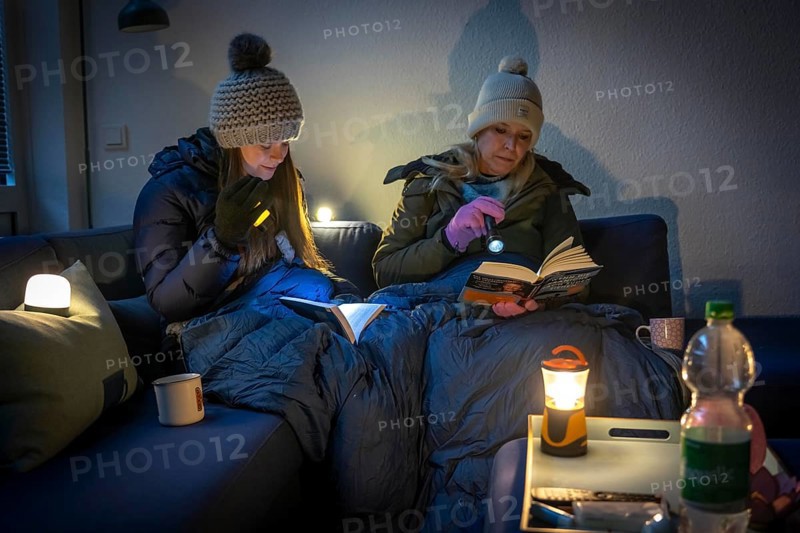 Symbolic picture blackout, extensive, prolonged power failure, in private surroundings, without electricity, heating, water, family on the sofa with warm winter clothes, sleeping bag, light from battery lights, battery lights, flashlights
