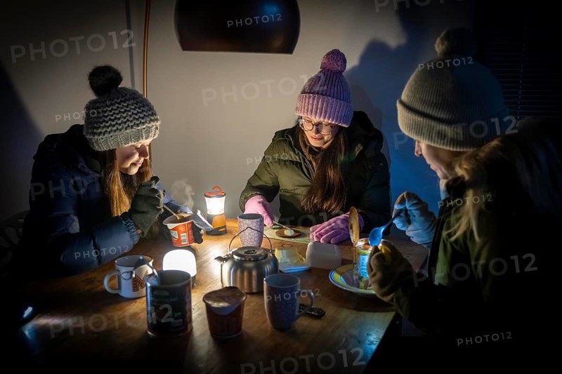 Symbolic blackout, extensive, prolonged power failure, in private surroundings, without electricity, heating, water, family at the dining table with warm winter clothes, sleeping bag, light from battery lights, flashlights, canned food, ready meals, heated on the camping stove, limited supply of food