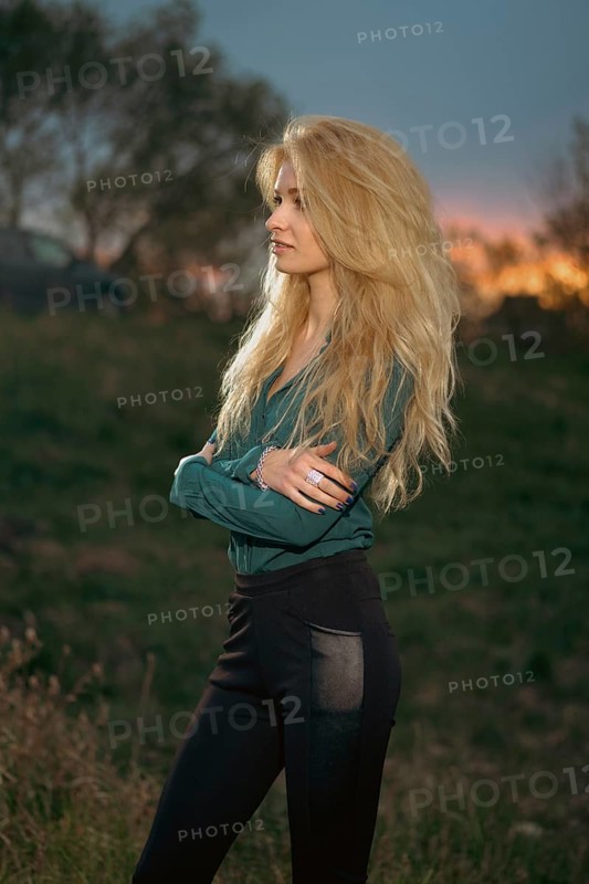 A woman stands with her arms crossed on a grassy hill. She faces the horizon where the sun is setting, creating a colorful sky. Trees and a vehicle are visible in the background