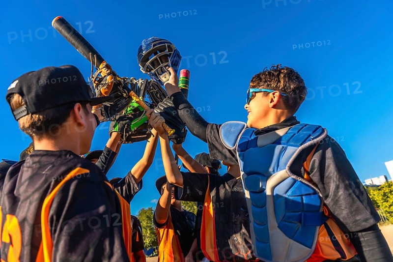 Young baseball players huddling together, lifting catcher's gear and a bat into the bright blue sky, celebrating a team achievement and showing unity on the field
