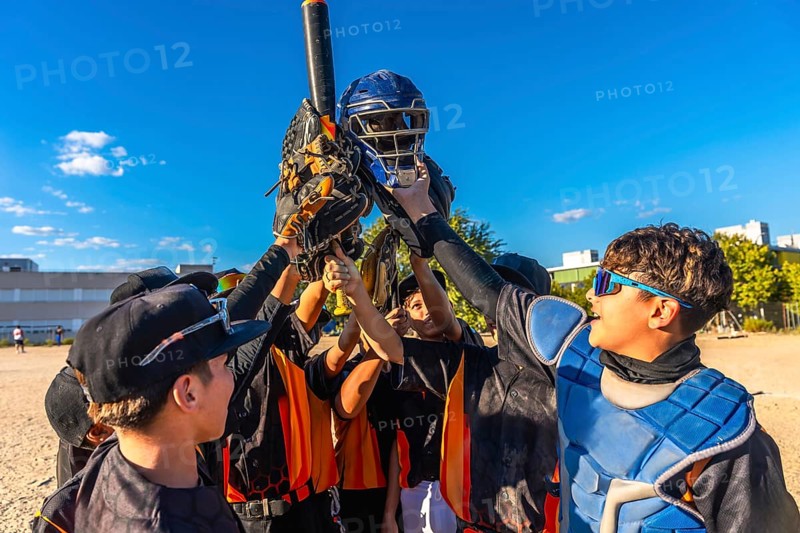 Baseball players huddling together, raising their catcher's helmet, bat, and gloves in the air, expressing team unity and triumph after a game on a sunny day