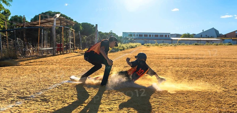 Two baseball players on a sunny field, one slides into a dusty base in a cloud of dirt while teammate watches the play, capturing speed, impact and competitive action