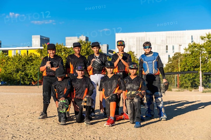 Young baseball players wearing uniforms and protective gear on a sunny day, standing and kneeling on a dirt field with trees and buildings in the background