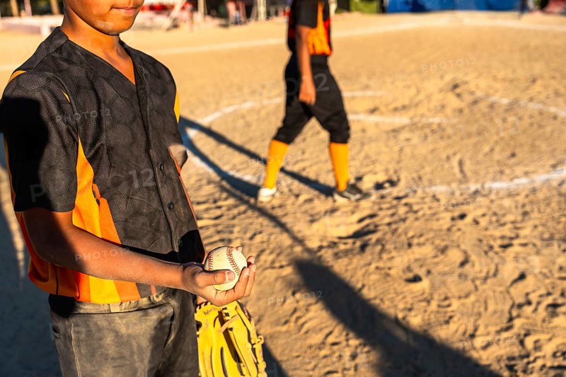 Young baseball boy in orange and black uniform holds glove and ball on sunlit dirt field during game, focused and ready while teammate stands blurred in background