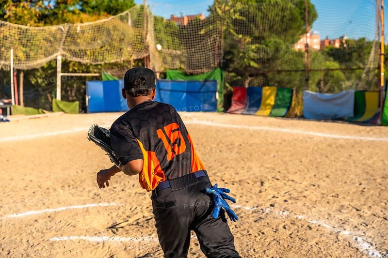 Player wearing a baseball uniform, cap, and fielding glove on a dirt field, throwing a ball during a game or practice session on a sunny day with a netting in the background