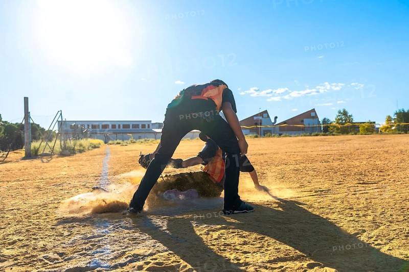 Two baseball players in action on a dirt field, with one player sliding headfirst into a base, creating a cloud of dust, while another player stands ready to make a play under a bright sunny sky