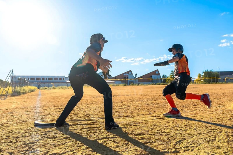 Youth baseball player sprinting hard across the dirt toward a base while coach or teammate awaits, showcasing speed, focus and summer youth sports action on a sunny field