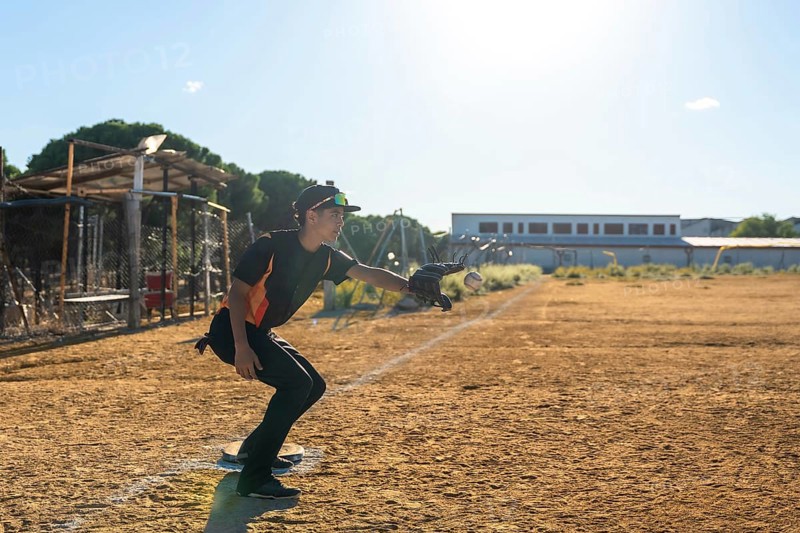 Baseball player in action on a dusty youth sports field, wearing uniform, cap and glove, extending his arm to catch a flying baseball under bright sunlight