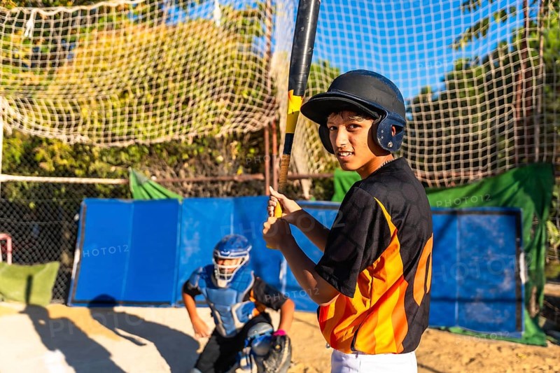 Young baseball batter wearing a helmet and looking at the camera while holding a bat, with a catcher in full gear positioned behind him on the field
