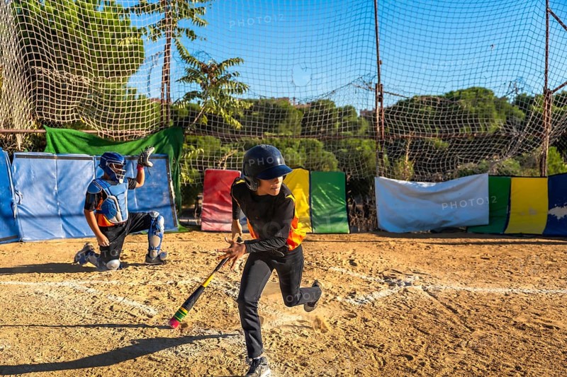 Kids playing baseball on a sunny day, an unidentifiable male batter wearing a helmet and looking back while running for first base and a catcher kneeling behind home plate