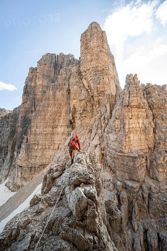 Mountaineer climbs on an exposed rock in the secured Via Ferrata Bocciere Centrale via ferrata, mountain landscape with spectacular rock towers in the back, Brenta-Adamello Nature Park, Trentino, Italy
