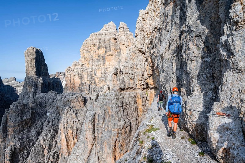 Two mountaineers on an exposed rock band in the secured Via Ferrata Bocciere Centrale via ferrata, spectacular mountain landscape with steep rocky peaks, Brenta Mountains, Parco Naturale Brenta-Adamello, Trentino, Italy