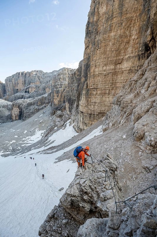 Mountaineer climbs on an exposed rock in the secured Via Ferrata Bocciere Centrale via ferrata, view of snow remains from the Vedretta del Sfulmini glacier, Brenta Mountains, Parco Naturale Brenta-Adamello, Trentino, Italy