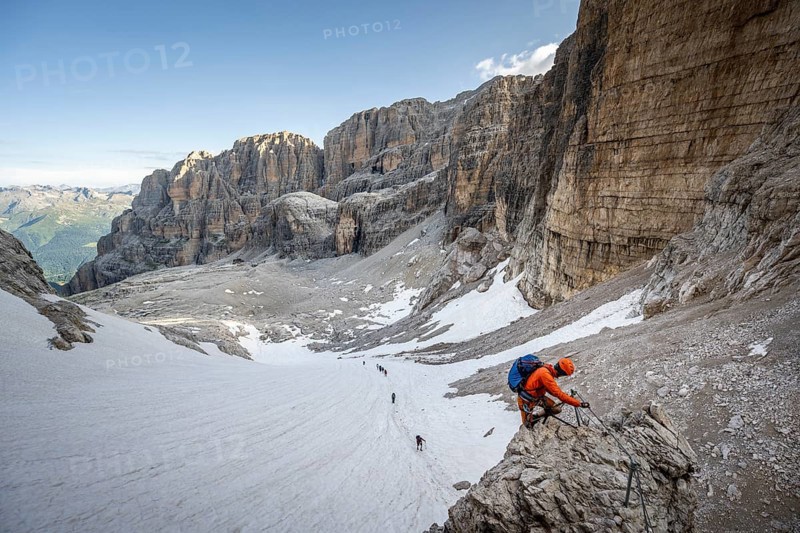 Mountaineer climbs on an exposed rock in the secured Via Ferrata Bocciere Centrale via ferrata, view of snow remains from the Vedretta del Sfulmini glacier, Brenta Mountains, Parco Naturale Brenta-Adamello, Trentino, Italy