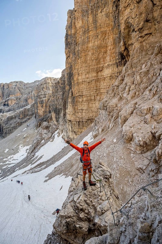 Mountaineer stretches his arms in the air, climbs an exposed rock in the secured Via Ferrata Bocciere Centrale via ferrata, view of remnants of snow from the Vedretta del Sfulmini glacier, Brenta-Adamello Nature Park, Trentino, Italy