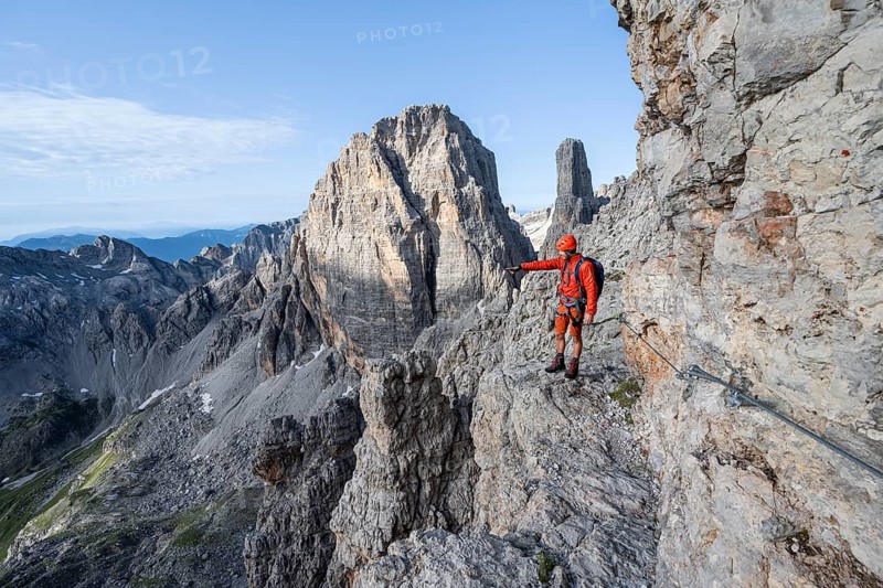 Mountaineer points into the distance, climbs an exposed rock band in the secured Via Ferrata Bocciere Centrale via ferrata, spectacular mountain landscape with steep rocky peaks, Brenta Mountains, Parco Naturale Brenta-Adamello, Trentino, Italy