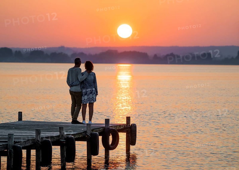 A couple stands on a jetty and looks at a colorful sunset over Lake Dümmer, Lembruch, Dümmer nature park Park, Lower Saxony, Germany