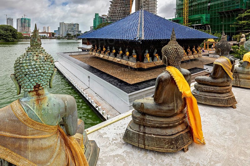 Buddha statues with yellow cloths sit in front of a modern cityscape on the water, The Seema Malaka Temple with the Buddha Statues in Colombo Sri Lanka