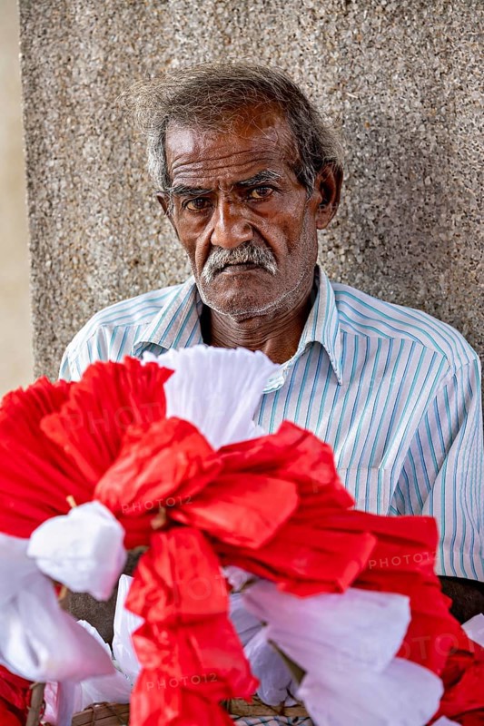 Close-up of an elderly man with red and white decorative flowers in the foreground, people making garlands in Colombo Sri Lanka City Hall