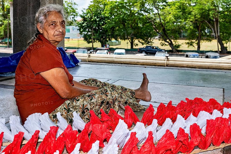 Elderly woman sitting outdoors working on red and white decorations, people making garlands in Colombo Sri Lanka City Hall