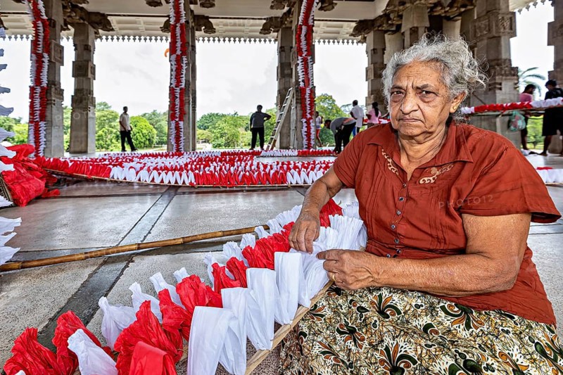Elderly woman sitting and making red and white decorations in a hall, people making garlands in Colombo Sri Lanka City Hall