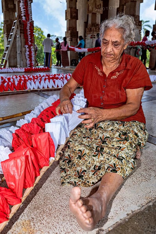 Elderly woman working with red and white decorations in an open hall, people making garlands in Colombo Sri Lanka City Hall