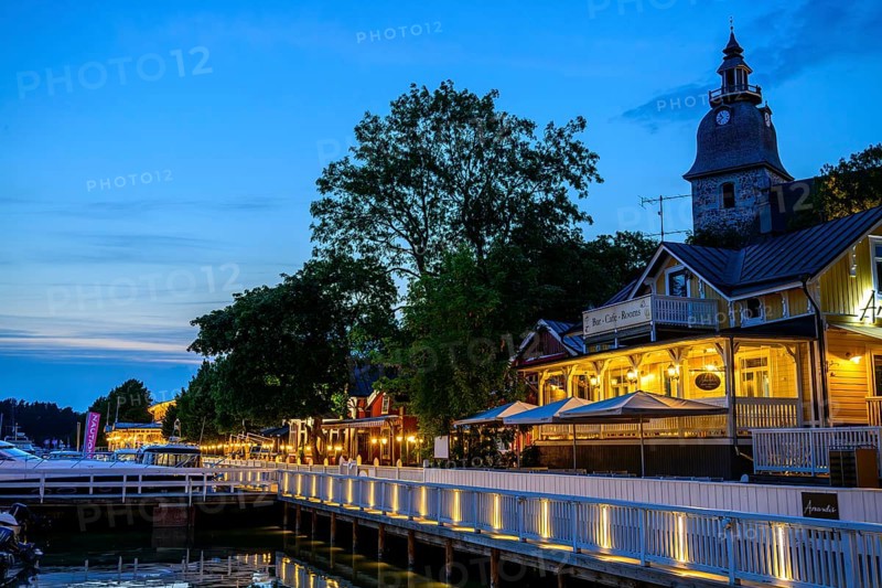 Street scene on Naantali harbour promenade, illuminated buildings and waterside boats at dusk, with church in the background, Naanatali, Varsinais-Suomi, Finland
