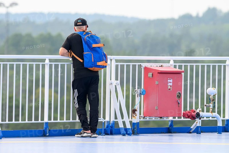 Man with blue backpack on deck of Finnlines ferry Naantali Kappelskär, overlooking wooded landscape, Aaland, Finland