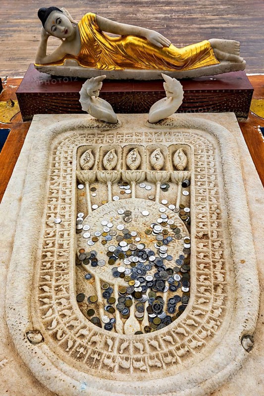 A reclining Buddha statue with gold robes above a carved stone surface filled with coins, symbolizing offerings in a spiritual setting, the Seema Malaka temple with the Buddha statues in Colombo Sri Lanka