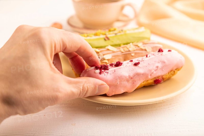 Glazed Eclairs on white wooden background and orange linen textile with hand, cup of coffee, side view, close up, selective focus