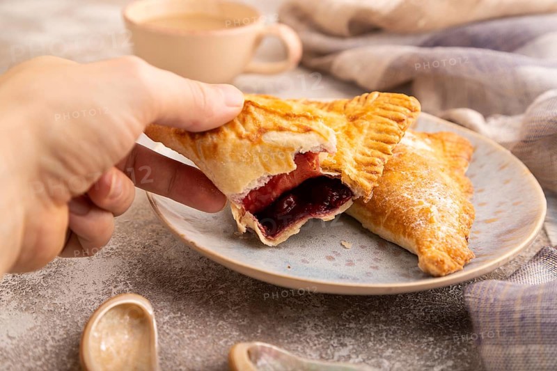 Triangles Pie, Puff Pastry, Samsa with cherry jam on brown concrete background and linen textile with hand, cup of coffee, side view, close up, selective focus