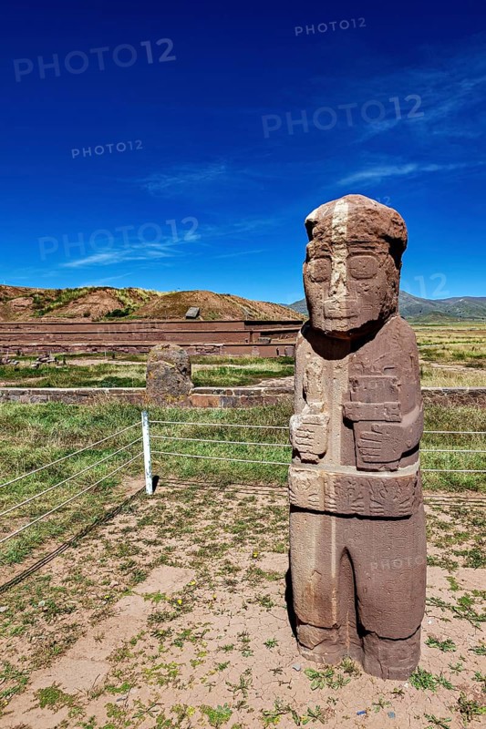Free-standing stone statue in a grass-covered ruined landscape under a blue sky, The archaeological site with the ruins of Tiwanaku in Bolivia
