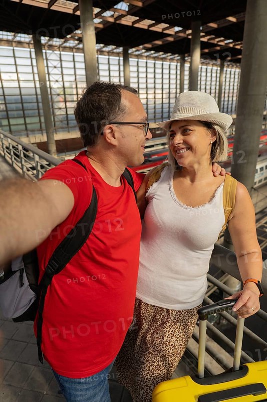 Smiling adult travel couple taking a selfie while looking at each other inside a modern train station with luggage. Romantic travel moment, connection, tourism and lifestyle concept